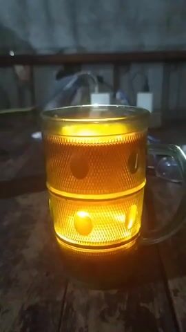 The video shows a close-up of a glass mug filled with a translucent amber liquid, possibly tea or beer. The mug is illuminated from within, casting a warm, golden glow through its textured surface, which features raised circular patterns. The mug has a handle and sits on a dark wooden surface, likely a table. In the background, out of focus, there are some electrical wires and power adapters. The lighting is dim, highlighting the illuminated mug as the central subject.