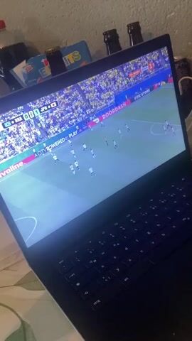 A laptop displays a soccer match, with the score showing BRA 0-0 JPN. The game is on a bright green field with a crowd in the stands. The laptop is on a surface covered with a patterned cloth, possibly a bedspread. To the left of the laptop, there are several bottles, including a dark soda bottle and some beer bottles. In the background, there is a light-colored wall. The scene is dimly lit, with the primary light source coming from the laptop screen.