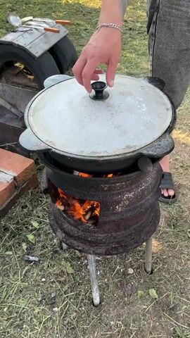 A person's hand, adorned with a bracelet, lifts the lid off a large, dark cauldron. The cauldron sits on a rustic, metal brazier fueled by an active fire with visible flames. Inside the cauldron, a stew-like dish is actively simmering, emitting a significant amount of steam. The dish contains visible ingredients such as chunks of vegetables like carrots and potatoes, along with what appears to be meat and possibly garlic. The setting is outdoors, with grass and scattered debris on the ground. In the background, there are elements that suggest a makeshift outdoor cooking setup, including what looks like a piece of discarded tire and some bricks. The person is wearing dark pants and sandals.