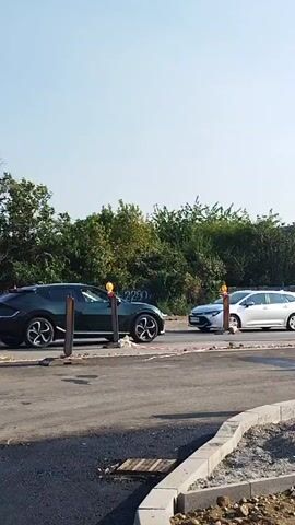 A dark green Kia EV6 is driving on a street. The street appears to be under construction, with piles of dirt and construction materials visible on the side. There are also temporary traffic barriers and signs. In the background, there are houses and a multi-story building. The sky is clear and blue.