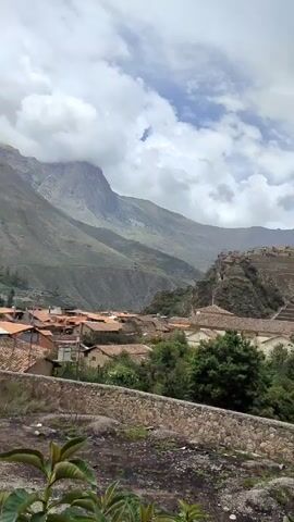 The video presents a scenic view of a mountainous landscape, possibly featuring agricultural terraces. The frame begins with a broader perspective, showcasing a mountainside with distinct terraced levels, likely ancient agricultural structures. In the foreground are buildings with clay-tiled roofs interspersed with greenery, and a stone wall that seems to demarcate the area. The camera then pans horizontally, revealing more of the structures below the terraces, and then focuses on a stone-paved path or street running alongside a lower stone wall. The sky is overcast with scattered clouds, lending the scene a muted, somewhat somber tone. The environment seems rural, marked by the presence of both natural and man-made features. There are no people visible in the video.