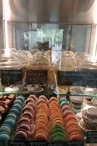The video shows a display case filled with various desserts. In the foreground, there are rows of colorful macarons in shades of blue, purple, orange, and green. Below the macarons, there are rows of chocolate-covered cakes and pastries, some with nuts and decorative drizzles of chocolate. Further back in the display case, there are other desserts, including what appear to be chocolate éclairs, tarts, and layered desserts in cups. Some of the desserts have handwritten labels indicating prices. The overall environment appears to be a bakery or patisserie.