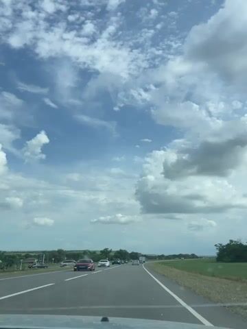 A first-person perspective video shows a car driving on a highway. The sky is blue with scattered white clouds. There are other cars on the road, both in front and in the opposite lane. The landscape on either side of the road is green and appears to be rural, with some trees and fields visible. The road has a white line marking the lanes. The car driving is moving at a moderate speed. There is a traffic sign visible on the right side of the road at 00:03.