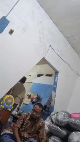 A man wearing glasses and a patterned shirt sits on the floor in a room. To his left is a yellow and blue fan on a wooden surface. To his right are several large, clear plastic-wrapped bundles. In the background, there is a doorway with blue and light-colored walls, and a small window with horizontal slats. The walls of the room are white, with some discoloration and cracking. The ceiling is white and slopes upwards.