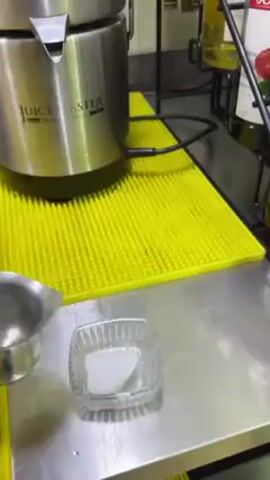 A hand is pouring water from a metal ladle into a small glass dish. The dish sits on a stainless steel surface next to a yellow, textured mat. In the dish is a white solid substance that, upon contact with the water, generates a cloud of white vapor. The vapor spreads out across the stainless steel surface and envelops the dish. To the left of the dish is a silver Juice Master juicer. In the background, bottles are partially visible on a wire rack.