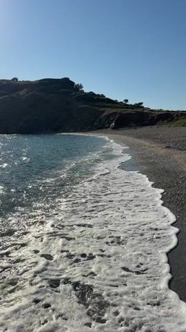 The video shows a beach with calm, blue water and small waves lapping at the shore. The beach is covered in pebbles, and a gentle slope of a hill rises behind it. The sky is clear and blue, and the overall scene is peaceful and serene. The camera is positioned close to the water, capturing the movement of the waves and the textures of the pebbles. The sunlight reflects off the water, creating a shimmering effect.