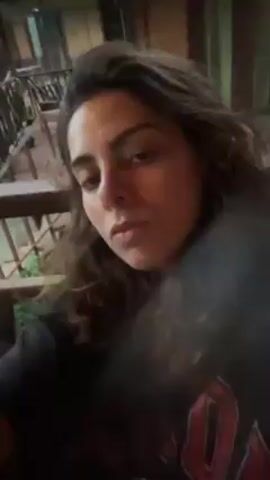 A woman with long dark hair and pale skin, wearing a black shirt with a red and white design, is sitting on a wooden porch. She is looking at the camera and talking. There is a smartphone and other objects scattered on the porch floor around her. Plants are visible through the porch railing in the background. The lighting is dim.