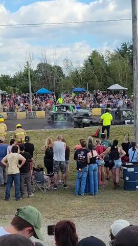 A demolition derby is underway. Spectators are visible in the background, some under tents. On the track, two cars are engaged in a collision. One car, painted green, is in the center of the frame, emitting smoke. Another car, partially obscured, is to the left. Several individuals in yellow vests appear to be directing the event. In the foreground, a group of people are standing and watching the derby, some with lawn chairs. The environment appears to be an outdoor event with a dirt track and grassy areas. The sky is partly cloudy.