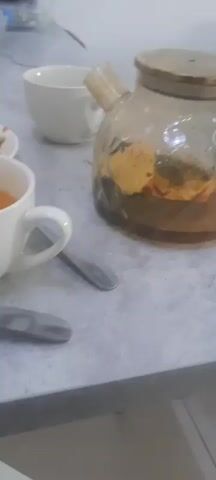 A close-up shot shows a table with a meal. In the foreground, two white teacups are visible, one filled with orange liquid. A glass teapot with a wooden lid sits to the right, also containing an orange liquid. Several plates with food are present, including a salad with tomatoes and cucumbers, and a plate of rice with meat and carrots. Spoons and forks are scattered around the table, suggesting a meal is in progress or has recently concluded. The table surface appears to be a grey, textured material. In the background, a plastic water bottle and some other objects are partially visible, suggesting an indoor setting. The lighting is dim.