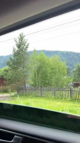 This media file is a video clip filmed from inside a vehicle, looking out of a window. The scene depicts a rural setting in Uznezya, Russia.

The primary subject is the landscape, characterized by a lush, forested mountain range in the background under a cloudy sky. In the mid-ground, there are several wooden fences, some enclosing yards with grass and scattered vegetation. There are also buildings visible, including what appears to be a small house with a green roof, a shed-like structure with a lattice facade, and other rustic structures. A solitary pine tree stands tall on the right side of the frame, near a wooden fence. A white utility pole with overhead wires is also present.

The activity taking place is movement through the landscape, as evidenced by the perspective from a moving vehicle and the shifting views. The time of day appears to be daytime, likely overcast due to the sky conditions. The weather seems to be mild, with no strong indications of rain or extreme temperatures. The overall mood is tranquil and serene, typical of a rural or mountainous environment.

There is visible text on a signpost on the right side of the frame, which appears to be a business sign. The text is in Russian and reads "НОЧЛЕГ" and a phone number "8-913-367-0567". "Ночлег" translates to "lodging" or "overnight stay," suggesting this is a place offering accommodation.

Other objects visible include a light-colored utility vehicle parked in a grassy area to the left, and a blue and white bus stop sign. The interior of the vehicle, specifically the dark-colored door panel and window frame, are visible in the foreground, framing the outdoor scenery.
