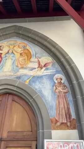 The video displays a close-up view of a fresco above a wooden arched doorway. The fresco depicts a religious scene with multiple figures. On the right side of the fresco, a man in a brown robe with a halo stands with his hands clasped. In the center and to the left, several figures are depicted in heavenly clouds. One figure, a woman in a blue robe with clasped hands and a halo, appears to be the central focus. She is flanked by two other figures, one in red and another in gold, also with halos. Two winged angelic figures, one on the left and one on the right, are blowing trumpets. The fresco is painted in vibrant colors against a blue sky background, with clouds and a dove motif at the top. The architectural elements of the building, including the stone archway and a red-beamed ceiling, are also visible. The lower portion of the video shows the top of a wooden double door. The overall scene is religious in nature, likely adorning the entrance of a church or similar religious building.