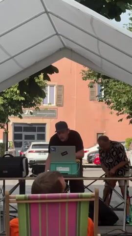A man wearing a black t-shirt and a baseball cap stands behind a table, looking down at a laptop. To his right, another man in a patterned shirt and shorts is bending over. The scene appears to be outdoors, possibly under a white tent. There are trees and buildings in the background.