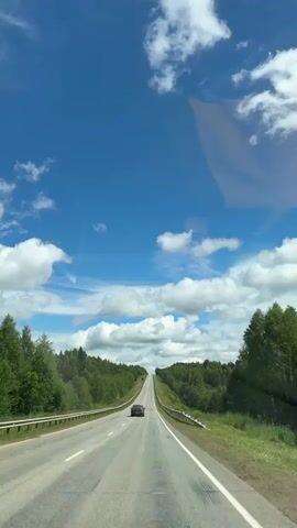 The video captures a view from inside a vehicle traveling on a highway. A dark-colored car is visible ahead on the road, moving away from the camera. The highway is flanked by lush green trees and dense forests on both sides. The sky is a vibrant blue, dotted with fluffy white clouds. The road appears to be in good condition with clear lane markings. The overall scene suggests a peaceful drive through a scenic, natural landscape on a clear day.