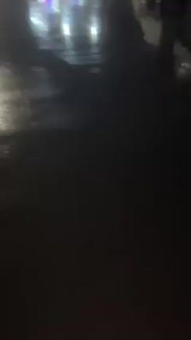 The video shows a dark and wet street at night. There are streetlights and the blurred lights of buildings in the background. The camera pans up to show overhead wires against the dark sky. It then focuses back down to the street where rain is falling heavily.