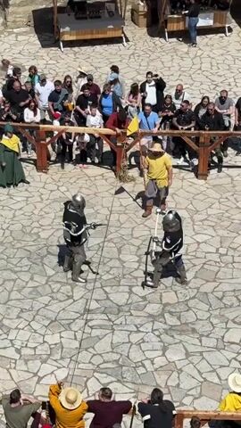 Two people dressed in medieval armor engage in a staged fight using poleaxes in a courtyard paved with flagstone. An umpire stands nearby holding a yellow flag and a staff. A crowd of onlookers stands behind a low wooden fence, observing the performance. The weather appears sunny.