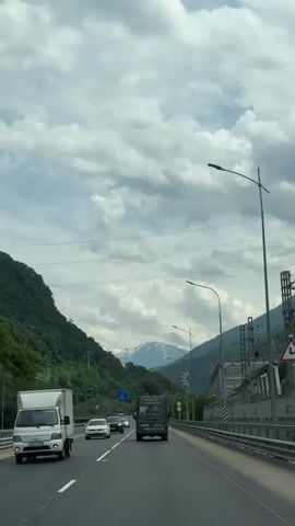 The video shows a view from a vehicle driving on a highway. The highway has multiple lanes and is surrounded by lush green mountains on the left and a more industrial area with bridges and structures on the right. The sky is filled with dramatic, heavy clouds, suggesting overcast or potentially stormy weather. Several vehicles are visible on the highway, including cars and a van. In the distance, snow-capped mountains can be seen peeking through the clouds. The overall impression is a scenic drive through a mountainous region under a cloudy sky.