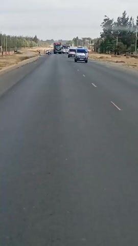 The video depicts a two-lane asphalt road with vehicles traveling in the same direction. The vehicles include a large semi-truck, several white vans, and a motorcycle. On the left side of the road, two individuals stand near a grassy area, and a motorcycle is parked nearby. The environment features a mix of natural and developed elements, including trees, grassy areas, power lines, and a building under construction. The sky is overcast, and the overall lighting suggests daylight conditions.