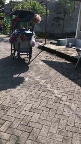 The video shows a food cart with a person preparing food. The cart is on a brick road with sunlight casting shadows. The cart is blue and has a canopy. There are bags of snacks and drinks on display. The person is wearing a headscarf and is in the process of serving a customer.