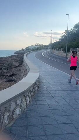 A person is running on a paved walkway next to a road. The walkway has a stone wall on the left side, separating it from the rocky coast and the sea. The sea is on the left side of the frame, with a faint horizon line visible under a pale blue sky with scattered clouds. The road curves to the right, with streetlights and trees lining the right side. The runner is wearing a bright pink tank top and black shorts, and is moving away from the camera. The overall scene is outdoors, likely during daylight or dusk.