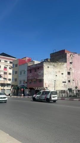 The video shows a street scene in what appears to be a city in Morocco, North Africa. A white taxi cab drives on a road in front of buildings of varying heights and colors, some of which are in disrepair. The sky is clear and blue. Other vehicles are parked or driving on the road. There are traffic lights visible.