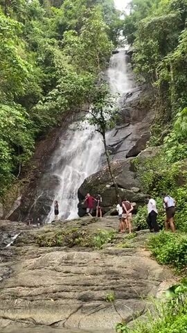 A long shot shows a multi-tiered waterfall dropping into a pool, framed by a dense green forest. Several people are visible at the base of the falls, scattered among the large gray rocks and lush vegetation. Some are wading in the shallow water, while others stand or walk on the rocky terrain. The lighting suggests a cloudy day.