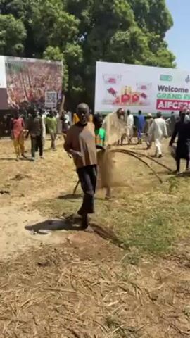 The video shows a group of people gathered on a dry, grassy field under a sunny sky. Many of the people are standing and walking around while carrying objects that appear to be fishing nets. There is a large banner in the background that reads "Welcome to the GRAND FINALE AIFCF 2026." Trees are visible in the background. The overall scene appears to depict some kind of outdoor event or competition related to fishing.