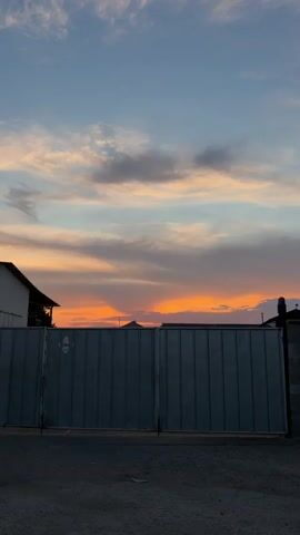 A vertical video shows a closed metal gate in front of a colorful sunset. The sky is a gradient of blue at the top, transitioning to orange and pink hues from the setting sun. Clouds are scattered across the sky, reflecting the sunset colors. To the left of the gate, a portion of a building with a white roof is visible. To the right, a cinder block structure is partially in view. The ground in front of the gate appears to be gravel or dirt. The gate itself is made of ribbed metal panels and is closed. There are no people or animals visible in the frame. The lighting suggests it is either sunrise or sunset.