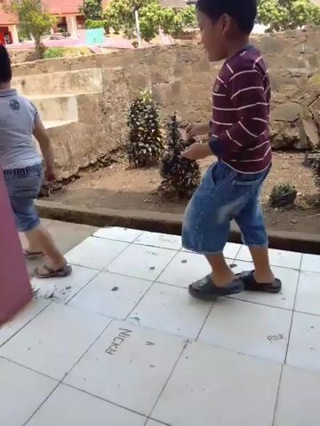 A boy in a striped shirt and blue shorts walks toward a set of concrete stairs. Another child, wearing a light blue shirt and denim shorts, is already walking up the stairs. The boy in the striped shirt follows, and both children ascend the stairs. The scene appears to be outdoors, with buildings and trees visible in the background. The ground is dirt and concrete, and there is a large pink pillar in the foreground.