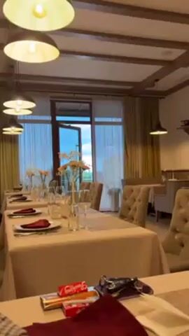 The video shows the interior of a dining room, likely in a restaurant or event venue. Tables are set for a formal meal, with beige tablecloths, wine glasses, plates, napkins, and floral centerpieces. The chairs are upholstered in a tufted beige fabric.  Large windows with sheer curtains let in natural light.  Outside the open door is a view of a building and greenery.  Near the camera, on one of the tables, are a pack of cigarettes, keys, and a wallet. The room is lit by hanging pendant lights. The camera slowly pans across the scene.  The overall impression is that the room is prepared for an event or a special occasion.