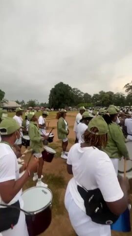 The video shows a group of people in a field. Most of the people are wearing green caps and green jackets with white shirts and shorts or skirts. Some of the people are holding drums and drumsticks. They appear to be part of a marching band or drumline. The field is covered in dirt and grass. There are trees and some buildings in the background. The sky is cloudy. The video is shaky, indicating it may have been filmed handheld.