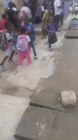 A group of children and adults are gathered outside a building, some of the children appear to be wearing school uniforms and carrying backpacks. The scene suggests a school drop-off or pick-up. The environment is an outdoor area with a paved surface, a building with a green roof, and a purple gate. There are vehicles visible in the background, indicating a street or road. The weather appears to be overcast. One child in the foreground is smiling at the camera.