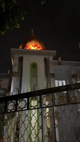 The video presents a static, low-angle shot of a mosque at night. The dominant feature is a striking golden dome adorned with red and yellow geometric patterns, illuminated brightly from within or by external lights. Below the dome, the upper part of a white building is visible, featuring ornate columns with intricate carvings and what appear to be vertical green decorative panels.

In the foreground, a dark, elaborate metal fence with repetitive star and interlocking geometric patterns partially obscures the view of the building. The fence itself has gold-colored accents or reflections. A few dark tree branches are subtly visible in the top-left corner against the dark night sky.

The scene is set at night, with the only visible light emanating from the building's illumination, highlighting its architectural details and casting a warm glow on the dome. There are no people visible, and no discernible activity is taking place other than the static view of the illuminated structure. No text is visible in the frame. The architectural style, particularly the dome and fence patterns, strongly suggests a mosque, consistent with the location context of Kaliwates, Indonesia.