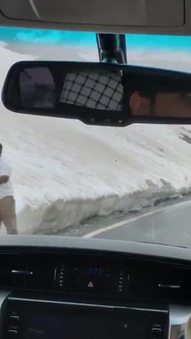 The video is taken from the interior of a car, looking forward through the windshield and reflecting in the rearview mirror. The road ahead is paved and curves to the right. On the left side of the road, there is a snowbank. Further down the road, there are three people standing near the snowbank, and a white SUV is visible approaching from the right. The time on the car's dashboard clock reads 12:27.