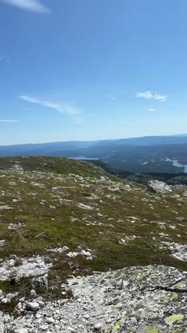 This video is a vertical, landscape view from a high elevation, likely a mountain top. The foreground is dominated by rocky, uneven terrain covered with sparse, dry grass and patches of moss or lichen. Large, weathered stones are scattered throughout. The middle ground shows a rolling landscape of green hills and valleys, dotted with numerous lakes of various sizes, reflecting the blue sky. Further in the distance, the terrain becomes more mountainous and hazy, with layers of blue-hued mountains receding into the horizon. The sky is a clear, bright blue with scattered white clouds, some of which are wispy and others more textured. The sun appears to be shining, casting light and shadows on the landscape. There are no people or man-made structures clearly visible in the frame.