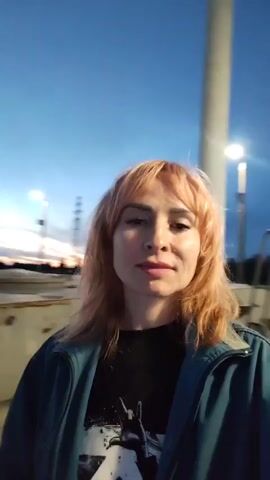 A woman with short, auburn hair and bangs, wearing a teal jacket over a black t-shirt with a graphic print, faces the camera. She has makeup on her eyes and lips and appears to be outdoors at dusk, with a blue sky above. In the background, there are lights and structures suggesting an urban or industrial setting. The video is a close-up shot of the woman's face, with slight movement as she looks around.