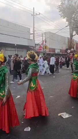 A street procession is underway. Several individuals are dressed in traditional cultural attire, including ornate headdresses, vibrant green and red outfits, and layered fabrics. Two women in the foreground wear these traditional costumes, one facing left and the other facing forward with her head turned to the side. In the background, a crowd of people lines the street, some watching the procession, others walking alongside. The street itself is paved and appears to have debris scattered across it. Buildings with awnings and signs are visible on either side of the street, suggesting an urban or town setting. Electrical wires crisscross overhead. The sky is overcast, with diffused light. The overall atmosphere suggests a cultural event or celebration.