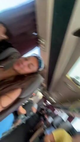 Two young people are sitting on a bus. The person in the foreground is a young woman with long brown hair, wearing sunglasses and a black top. She is smiling at the camera. The person behind her is a young person with short brown hair, wearing a purple t-shirt. They have their head resting on their hand and are looking away from the camera, with earbuds in their ears. The bus has red curtains on the windows, and trees and sky can be seen through the windows, indicating the bus is in motion.
