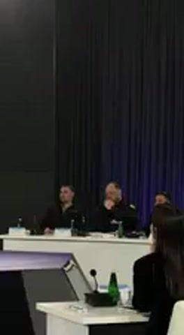 The video shows an indoor event, possibly a conference or meeting, set in Barvinok, Ukraine. In the mid-ground, two men are seated side-by-side at a long white table. Both are wearing dark, possibly black, jackets. The man on the left has dark hair and is looking forward, appearing attentive. The man on the right, with a beard, is leaning slightly forward with his right hand resting on his chin, suggesting deep thought or listening intently.

On the white table in front of them, there are several objects including microphones, small green bottles (likely water), and papers. In the foreground, the back of a woman's head and shoulders is visible, also seated at a table which holds a laptop and another microphone. The background consists of dark, floor-length curtains, possibly blue or black, giving a formal appearance to the setting. The lighting is artificial, illuminating the speakers and the table area. The overall atmosphere suggests a focused and formal discussion or presentation. Audio indicates the discussion is in Ukrainian, mentioning a "youth congress" (молодіжний конгрес) and the "development of the country" (розвитку країни), suggesting topics related to youth and national progress. No specific text is clearly legible on any visible papers or screens.