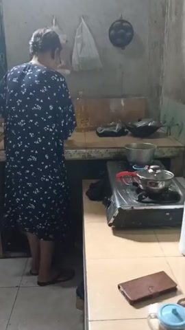 A woman wearing a dark blue dress with white floral patterns stands in a kitchen, facing away from the camera. She is positioned in front of a tiled countertop, with a stove to her right. On the stove, there is a pot with a lid, and a burner is lit, producing a blue flame. Another pot is on a shelf above the stove. A bottle of cooking oil and other items are on the countertop. The kitchen appears to be a functional space with tiled walls and surfaces.