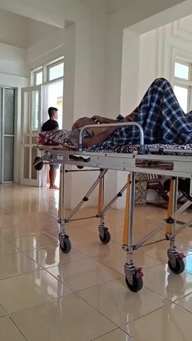 A patient is seen lying on a gurney in a well-lit room, covered with blankets. The gurney is silver with yellow accents and black wheels. The room has light-colored tiled floors and white walls with multiple windows, which let in ample sunlight. Through an open door, one can see a person standing outside and an ambulance parked in the distance. The overall atmosphere suggests a medical or care facility.