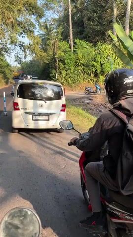 A person wearing a black helmet and backpack rides a red and black motorcycle on a paved road. The motorcycle has the word "Supra" visible on its side. The rider is wearing a dark jacket and grey pants. To the left of the motorcycle, a white van is parked. The road is bordered by lush green trees and foliage. The sky is bright, suggesting it is daytime. In the background, another blue motorcycle is visible. The scene appears to be in a rural or tropical environment.
