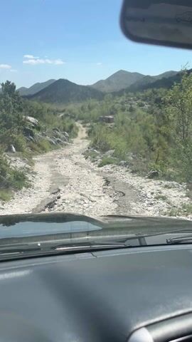 The video shows a first-person view from inside a car driving on a rough, rocky road. The landscape is mountainous, with sparse green vegetation and a clear blue sky above. The road is unpaved and appears quite challenging due to the numerous rocks and uneven terrain. The car's dashboard is visible at the bottom of the frame, along with a portion of the hood and the rearview mirror. The general impression is of an off-road driving experience through a remote, natural environment.