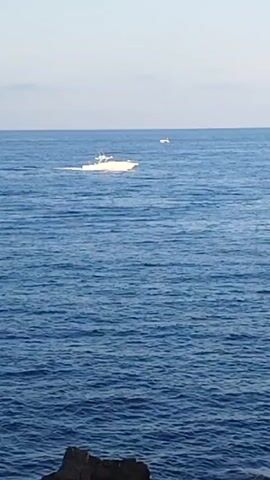 A white boat with a dark hull travels across a calm blue ocean, leaving a visible wake. The boat is moving from left to right across the frame. In the distance, another smaller white object, possibly another boat or debris, is visible. The water is deep blue and shows small ripples. The horizon is visible in the background, with a clear sky above. In the foreground, dark rocks are visible along the bottom edge of the frame, suggesting a rocky shoreline or outcropping. The lighting suggests it is daytime, with soft sunlight.