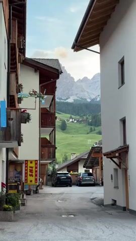 The video shows a view down a narrow street between two buildings. The street is paved and there are two cars parked on the right side. The buildings on either side are made of wood and stone, and have balconies with flower boxes. In the background, there are green hills with scattered houses and trees, and in the distance, there are tall, snow-capped mountains. The sky is overcast.