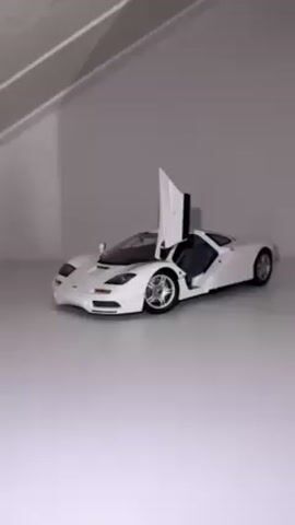 A static shot shows a white McLaren F1 model car on a white surface. One door of the model car is open. The car is positioned against a white background with a shadow cast from above.