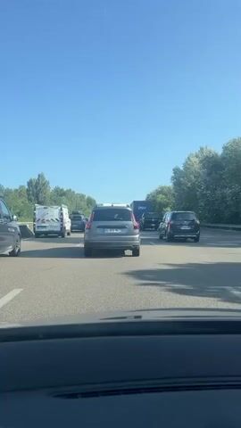 This is a point-of-view video taken from inside a vehicle, looking forward through the windshield. The vehicle is stopped in heavy traffic on a highway. Several cars and vans are visible in front, as well as a large truck in the distance. The road is paved, and there are trees and foliage on the right side of the highway. The sky is clear and blue, indicating a sunny day. The time stamp reads 00:00 to 00:05.