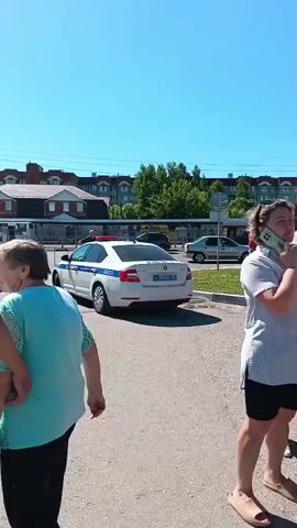 A white and blue police car is parked on a paved lot in front of a bus stop with a few people standing in the background. A woman in a light blue top and black pants is walking in the foreground. A man in a white shirt and dark pants is walking beside her. The sky is bright blue and clear.