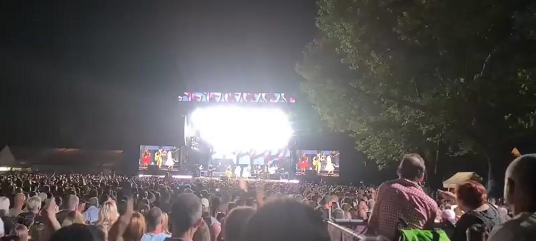 A wide-angle view of an outdoor concert at night, looking towards a brightly lit stage. The stage has a large screen displaying a colorful abstract pattern above the performers, and two smaller screens on either side showing close-ups of performers. There are multiple stage lights illuminating the area. A large crowd of people is visible in the foreground and midground, facing the stage. Most people are standing, and some have their arms raised. The environment appears to be a park or open field, with trees visible in the background on the right side of the frame. The overall atmosphere is one of an energetic performance.