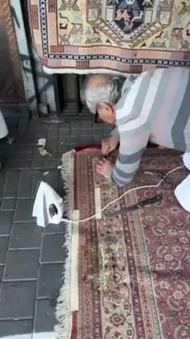 The video depicts an older man kneeling on a patterned rug on a sidewalk and repairing it. The man has white hair and is wearing glasses and a striped sweater. He is focused on his work, manipulating the fabric of the rug with his hands. An iron and a machete are lying on the rug next to him. Above him, a rug is hanging. The sidewalk is made of gray, square tiles. A metal grate can be seen in the background.