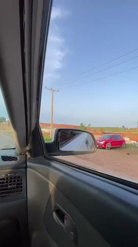 The video shows a car driving down a dirt road in what appears to be a developing area. The view is from inside the vehicle, focusing on the passenger side window and side mirror. Outside, there are houses under construction, utility poles, and a clear blue sky. The road is unpaved, and the surrounding land looks bare and somewhat dusty.  The car is moving at a moderate speed, passing by the developing neighborhood.  The side mirror reflects the ongoing scenery, including more houses and the red shirt of the person filming. The interior of the car is visible, revealing a slightly worn dashboard.